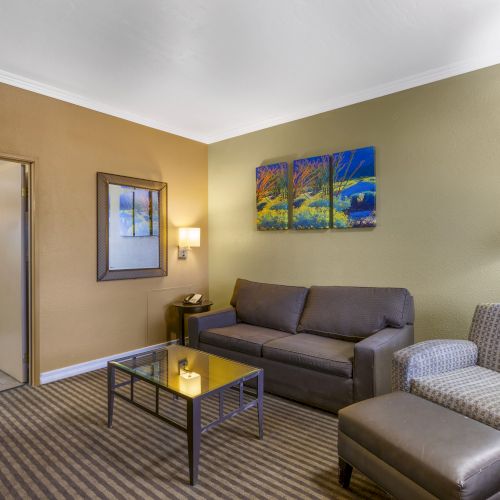 The image shows a cozy hotel room with a sofa, armchair, coffee table, and art on the walls, leading to a bedroom with a bed.