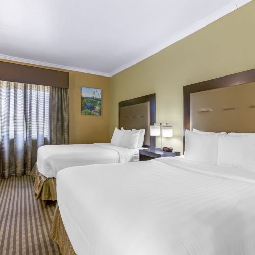 The image shows a hotel room with two neatly made beds, a work desk and chair near a window, and a painting on a beige wall.