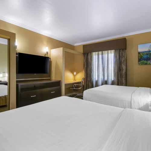 A hotel room with two double beds, a wall-mounted TV, desk, and a large mirror, decorated in neutral tones with a window letting in natural light.