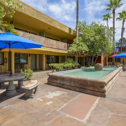 A courtyard featuring benches, tables with blue umbrellas, and a central water fountain surrounded by trees and a two-story building.