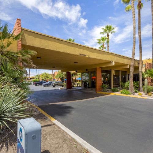 The image shows the entrance of a Best Western Royal Sun hotel with a covered driveway, surrounded by palm trees, plants, and a clear sky.