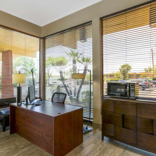 The image shows a small office or waiting area with a desk, chairs, microwave, coffee maker, and large windows with blinds, overlooking the outside.