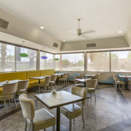 A brightly lit diner with several tables and chairs, modern light fixtures, yellow bench seating, large windows, and whimsical wall art.