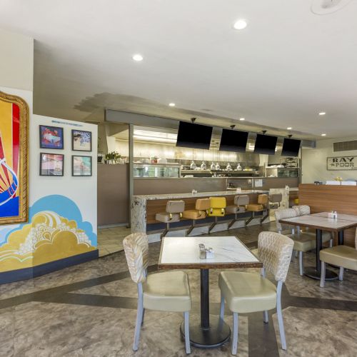 A modern diner interior with seating, wall art featuring a retro-style figure, overhead lighting, and a view of the open kitchen and counter area.