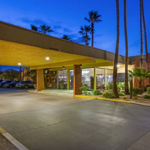 The image shows the entrance of a Best Western hotel with a well-lit sign and parking area, surrounded by palm trees and evening sky.