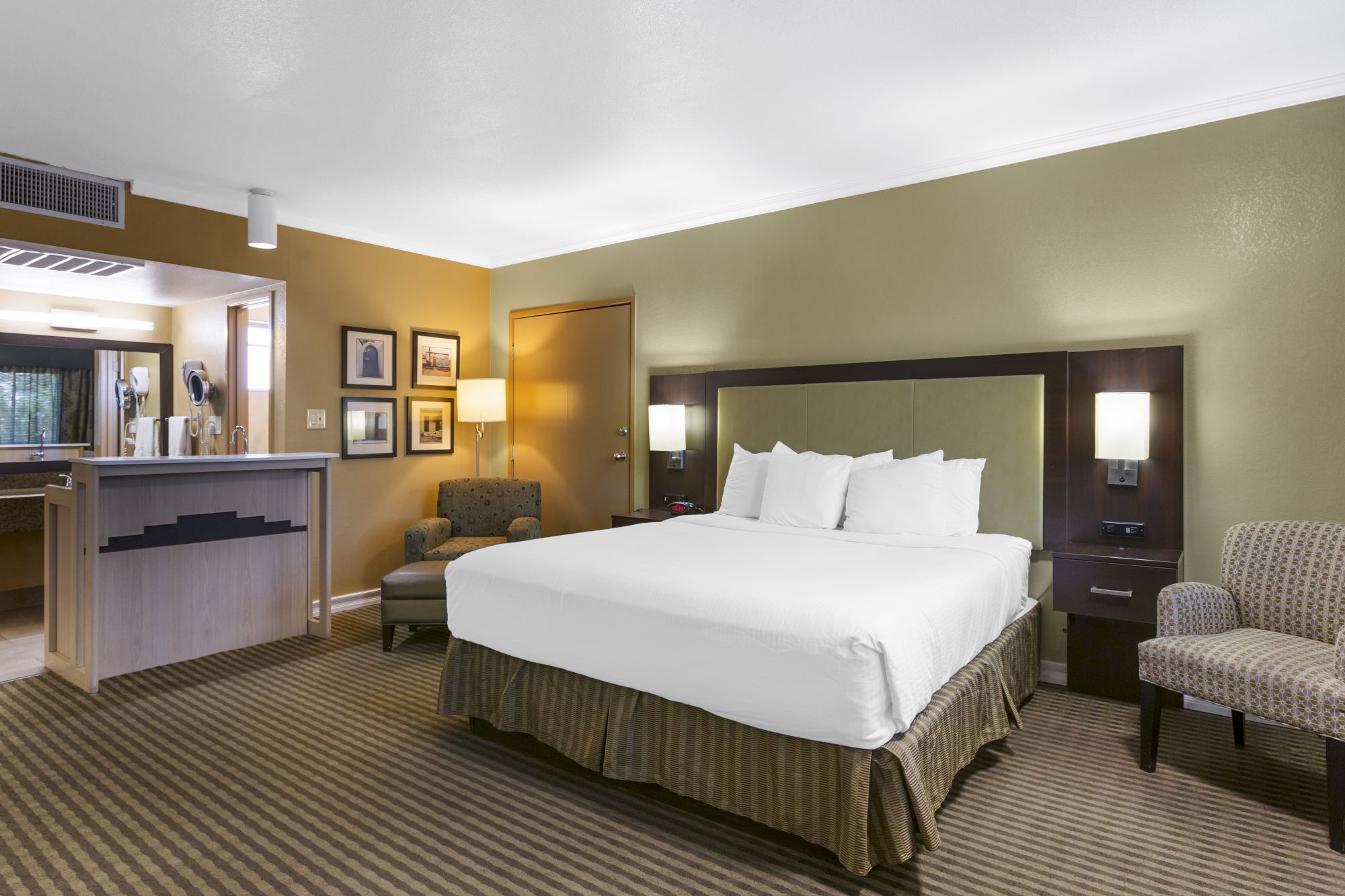 The image shows a hotel room with a king-sized bed, two chairs, a small table, a kitchenette, framed pictures, and warm lighting.