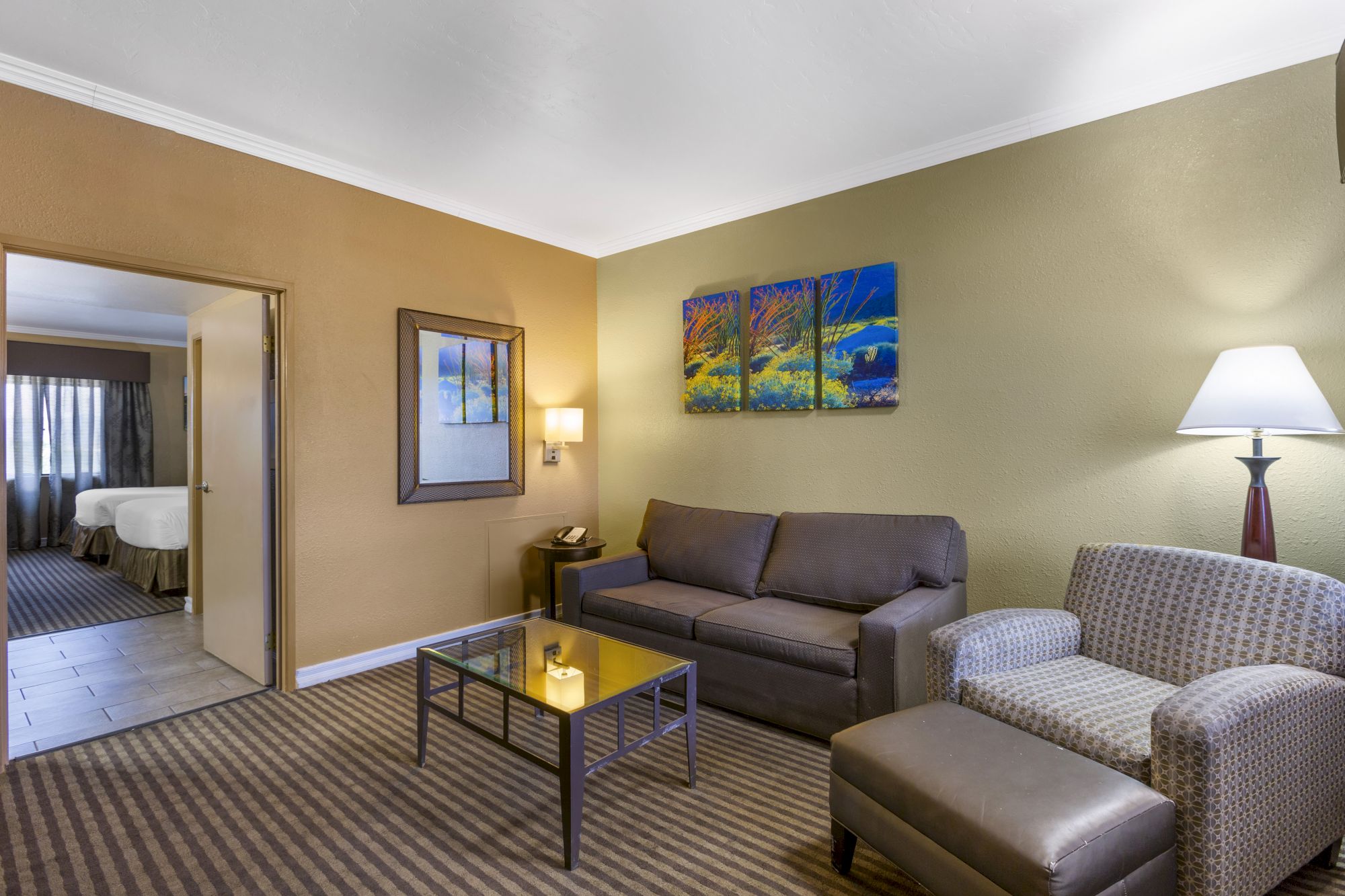 The image shows a living room with a loveseat, armchair, coffee table, lamp, and paintings on the wall. A bedroom is visible through the doorway.