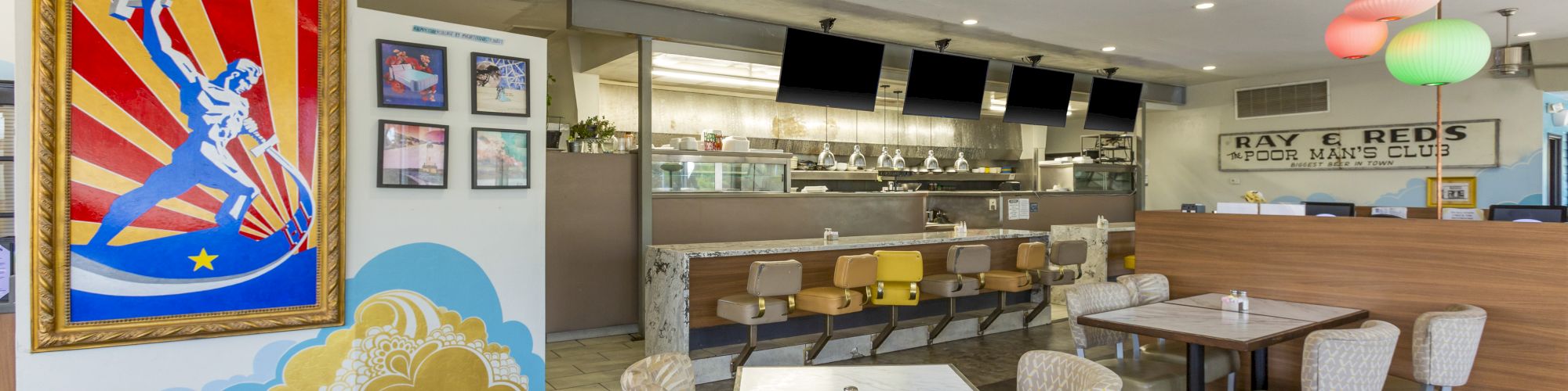 A retro-style diner interior with various seating arrangements, colorful wall art, and a counter area with stools and hanging pendant lights in the background.