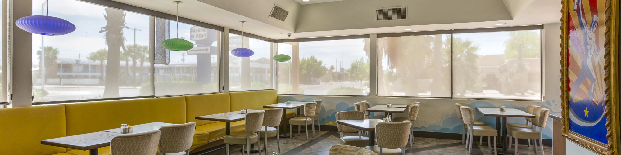 The image shows an empty restaurant interior with yellow booths, various chairs, and tables, large windows, and a colorful mural on the right wall.