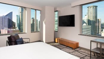 A modern hotel room with a large bed, armchair, wall-mounted TV, and windows offering a cityscape view of tall buildings and a blue sky.
