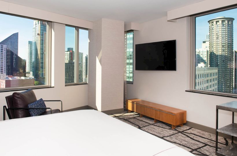 A modern hotel room with a large bed, armchair, wall-mounted TV, and windows offering a cityscape view of tall buildings and a blue sky.