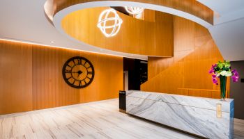 A modern lobby area with a marble reception desk, decorative wooden elements, a large wall clock, and floral arrangements on the desk.