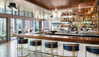 A modern bar with a wooden counter, high black and gold stools, a well-stocked shelf of liquor, and large windows overlooking city buildings.