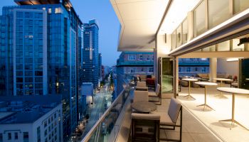 A modern rooftop patio with seating areas and tables overlooks a city street with tall buildings in the evening.