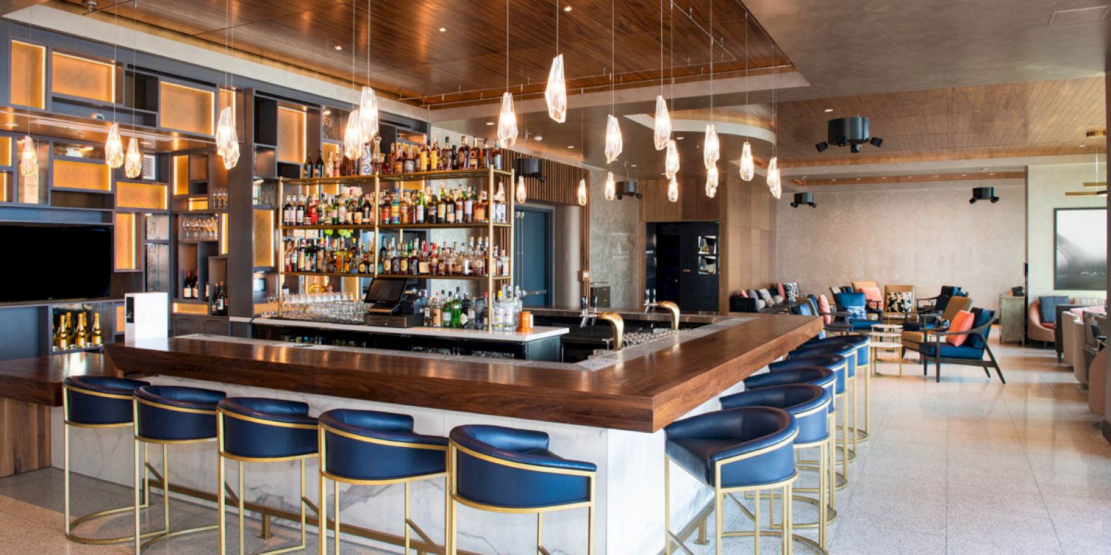 This image shows a modern bar with blue seating, stylish lighting, and a fully stocked bar counter, adjacent to a lounge area with plush seating.