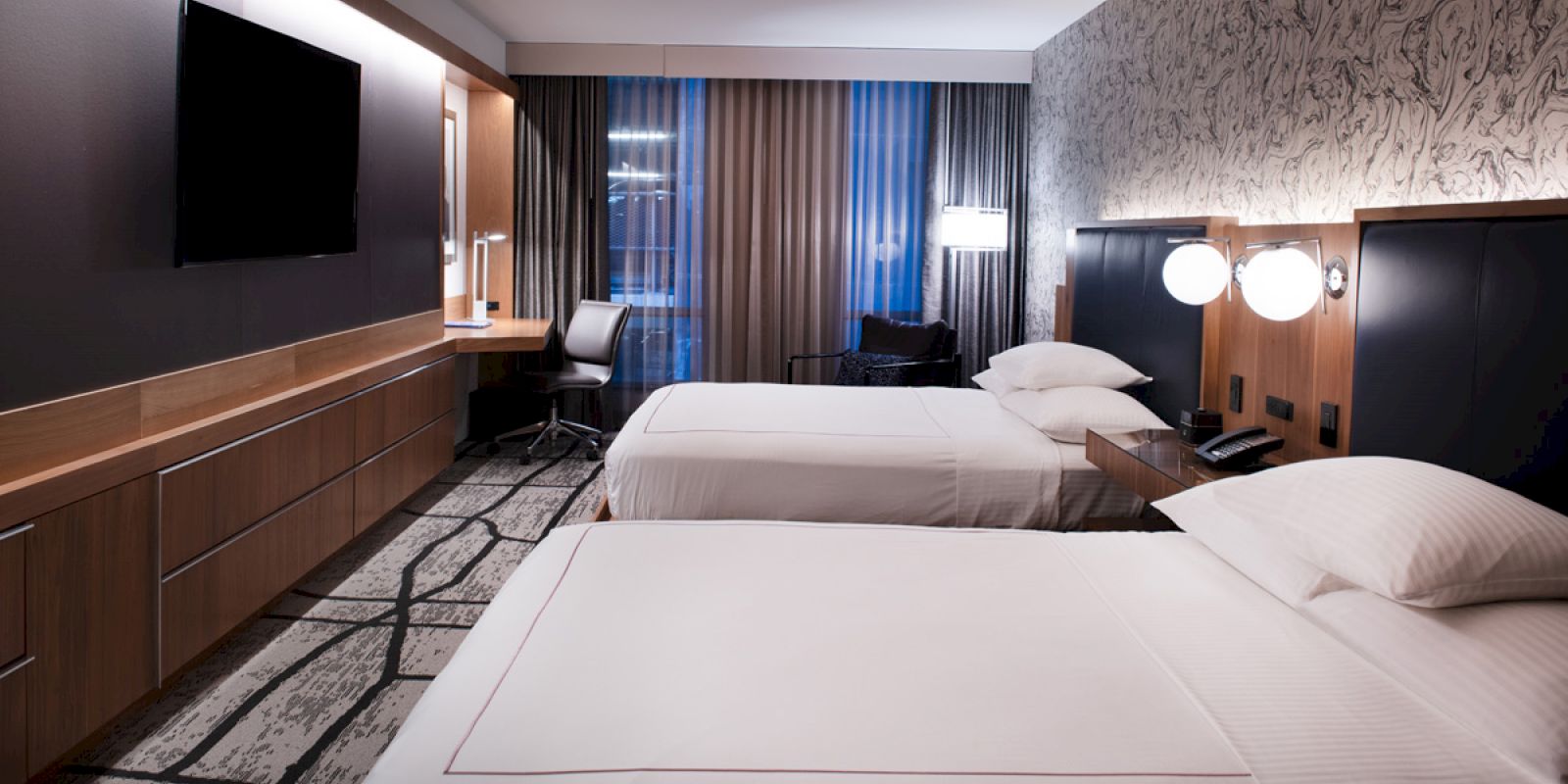 A modern hotel room with two neatly made beds, a desk with a chair, a wall-mounted TV, and stylish decor and lighting, appears well-lit.