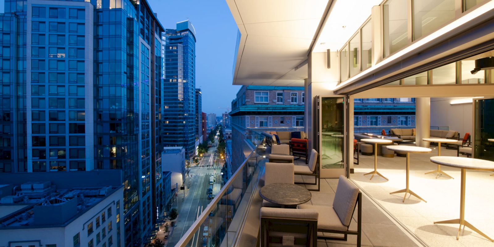 The image shows a modern cityscape viewed from a balcony at dusk. The balcony has seating and tables, with tall buildings in the background.