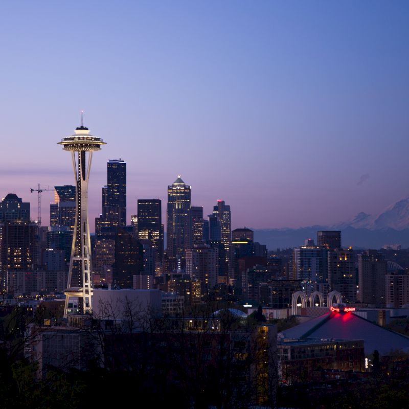 The image shows the Seattle skyline at dusk, featuring the Space Needle and Mount Rainier in the background, with city lights starting to illuminate.