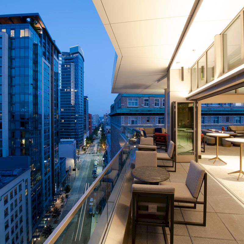 A modern balcony with chairs and tables overlooks a city street lined with tall buildings at twilight, showcasing an urban architectural setting.