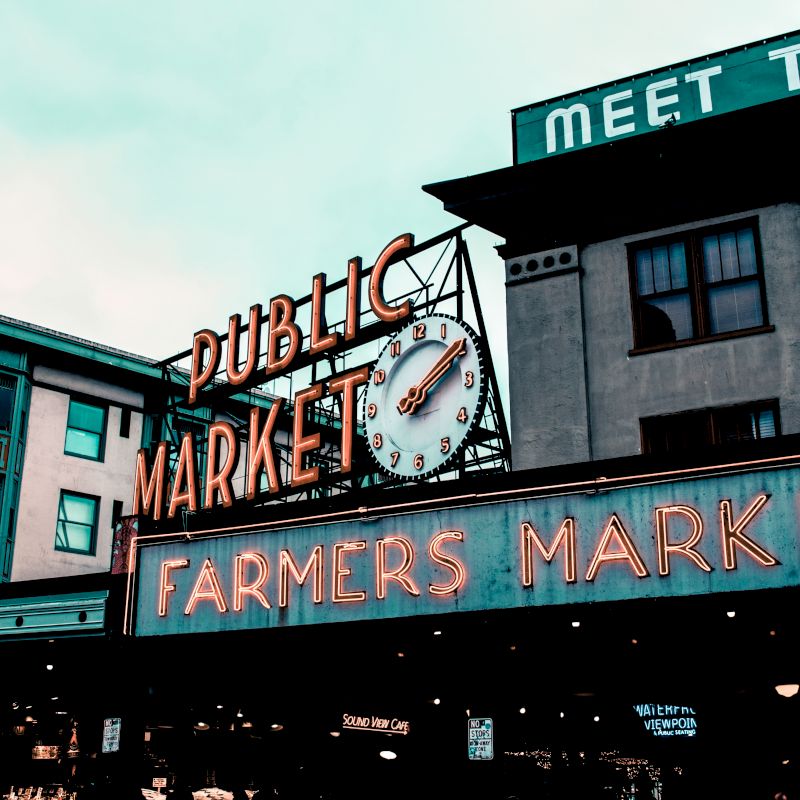 The image shows the front of a farmers market with a neon sign reading "PUBLIC MARKET" and "FARMERS MARKET" along with a clock.