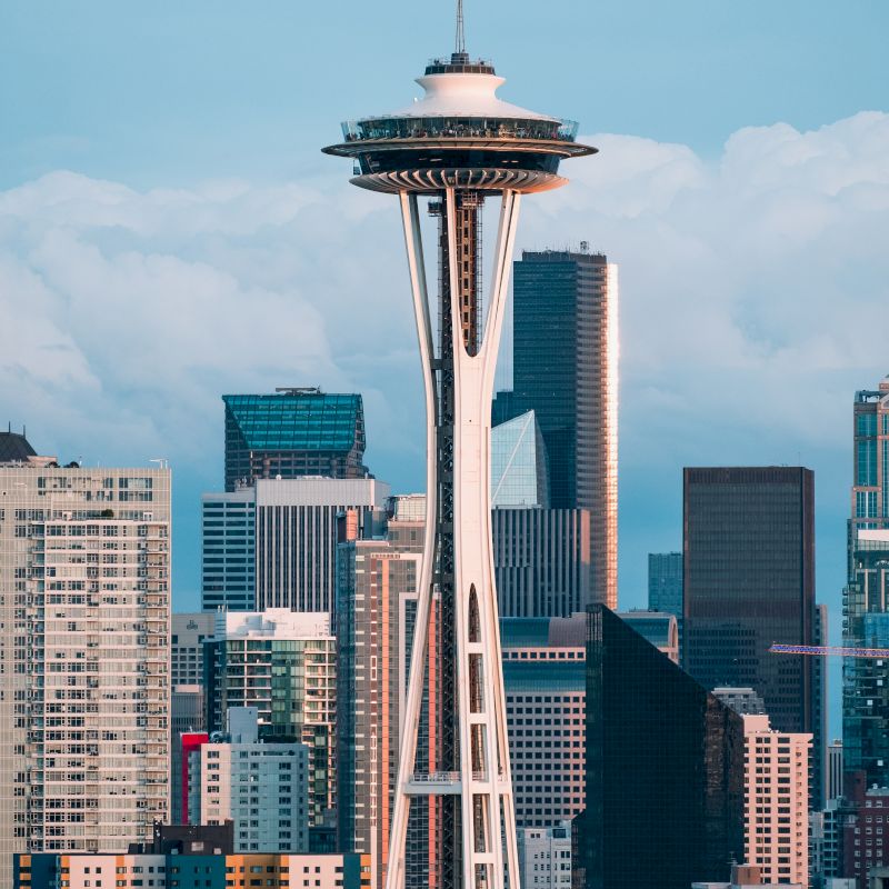 The image features the Space Needle against a backdrop of skyscrapers in a cityscape, under a clear sky with some clouds in the background.