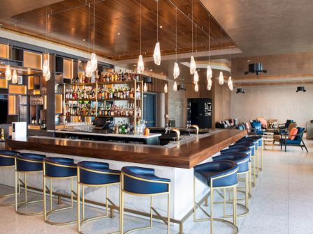 This image shows a modern bar with a long wooden counter, blue barstools, hanging lights, and a well-stocked shelf of liquor bottles, located in a spacious room.