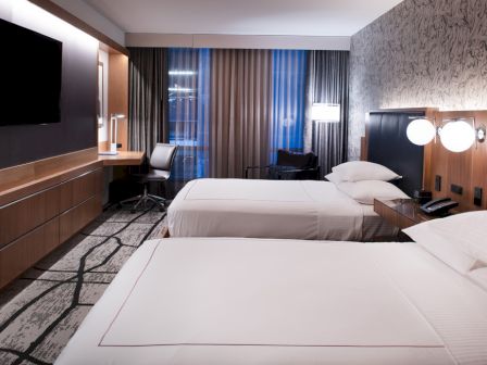 A modern hotel room with two neatly made beds, a flat-screen TV, a desk with a chair, and contemporary decor, featuring patterns and subtle lighting.