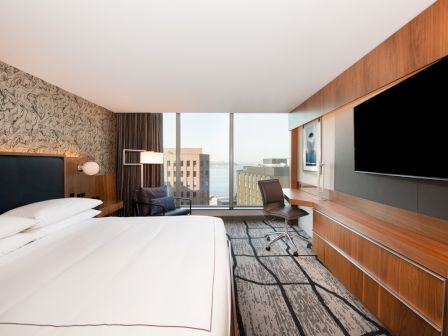 A modern hotel room with a large bed, a desk with a chair, a wall-mounted TV, and a window with a city view.