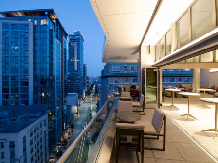 A modern outdoor balcony in a high-rise building with tables and chairs, overlooking a cityscape with tall buildings at dusk.