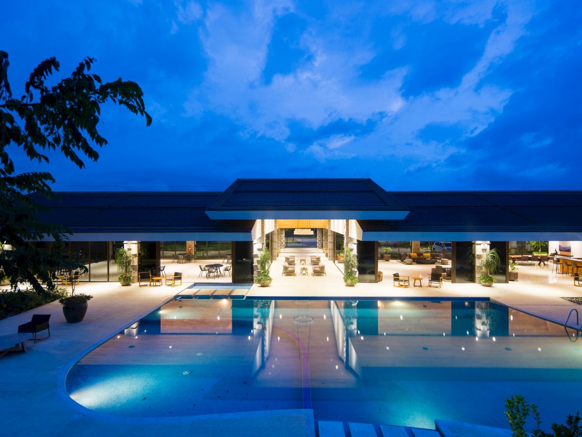 An illuminated outdoor pool area at night, with lounge seating and a modern building in the background under a partly cloudy sky.