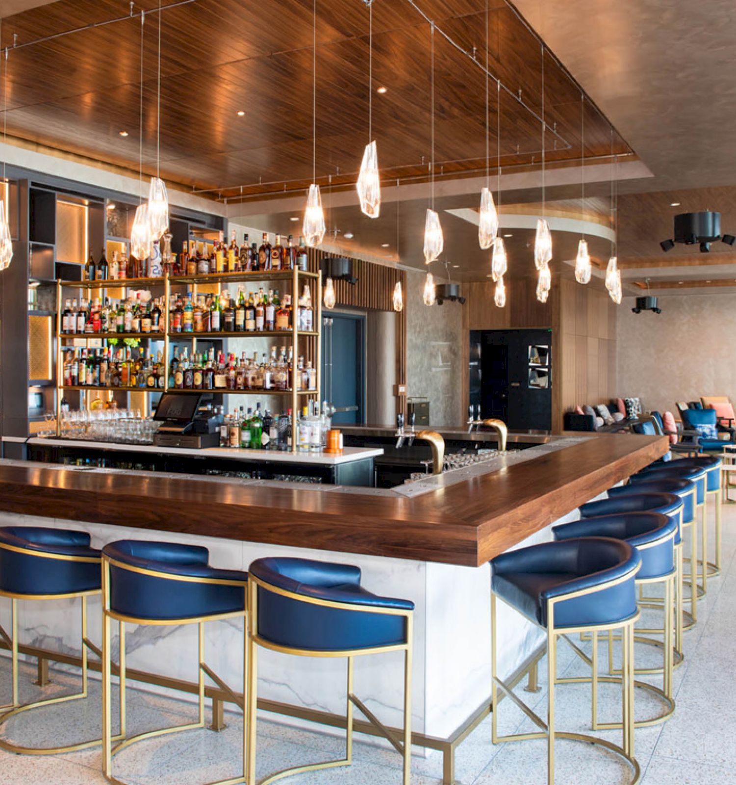 The image shows a modern bar with a well-stocked liquor shelf, blue barstools with gold frames, pendant lighting, and a seating area in the background.
