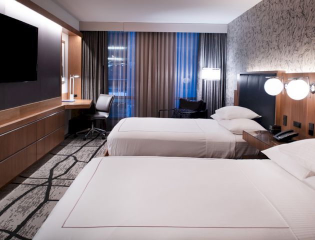 The image shows a modern hotel room with two double beds, a TV on the wall, a work desk and chair, a lamp, and floor-to-ceiling windows.