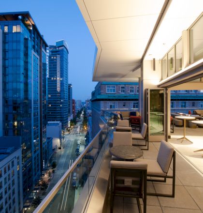 A modern cityscape at dusk with high-rise buildings and a balcony featuring tables and chairs overlooking the view.