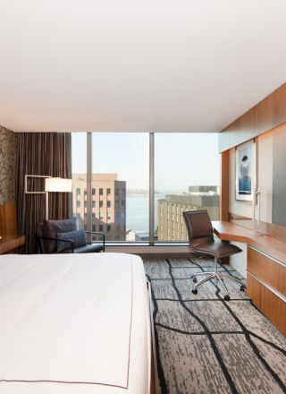 The image shows a modern hotel room with a large bed, a desk with a chair, a flat-screen TV, and a view of city buildings through a big window.