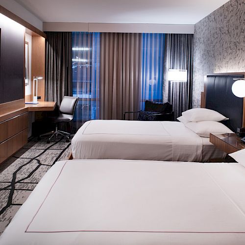 Modern hotel room with two beds, a TV, desk, and chair. The room features contemporary furniture, wall art, and a large window with curtains.