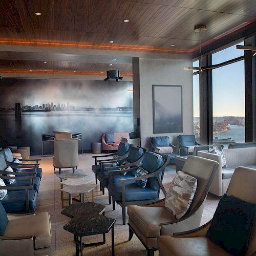 A modern, spacious lounge with multiple seating options and large windows offering a waterfront view. The city skyline mural adds ambiance.