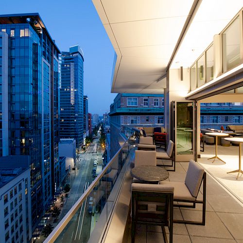 A modern balcony with seating overlooks a city street surrounded by tall buildings during the evening, with lights illuminating the scene.