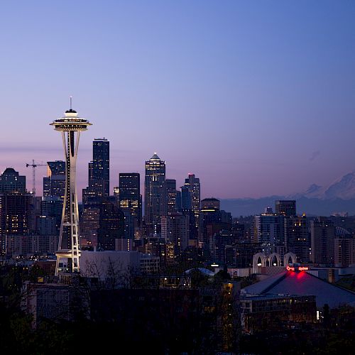 This image shows the skyline of Seattle at dusk, featuring the Space Needle, numerous buildings, and Mount Rainier in the background.