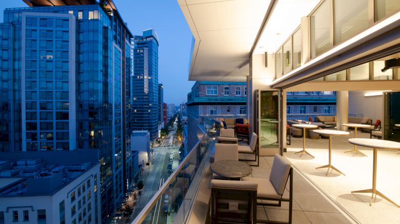 The image shows a modern rooftop balcony overlooking a city street with tall buildings, featuring tables, chairs, and floor-to-ceiling windows ending the sentence.