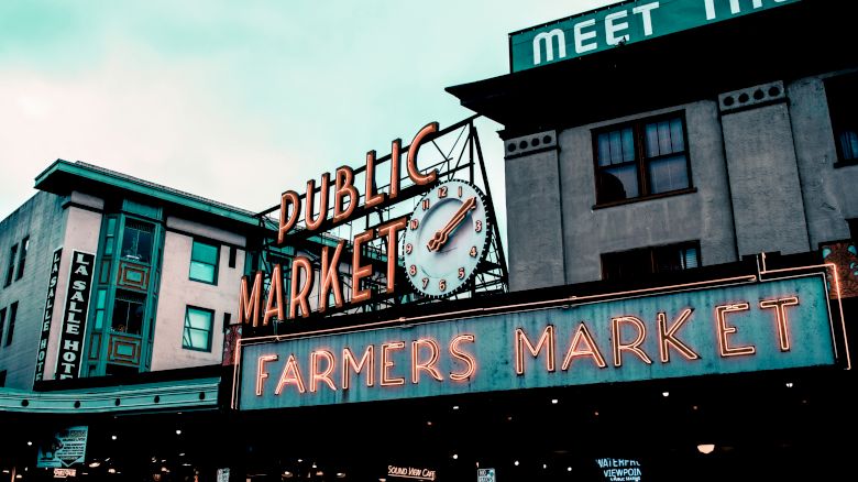 A neon sign for a public market and farmers market, along with a clock, is shown in front of an old building.