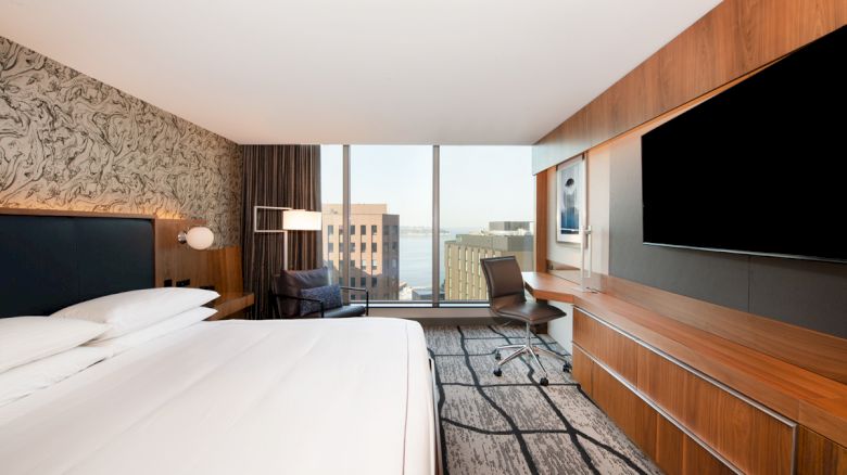 A modern hotel room with a large bed, desk, chair, TV, and window with a cityscape view. The decor is contemporary with wood accents.