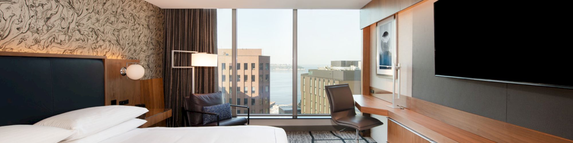 A modern hotel room with a large bed, flat-screen TV, desk, chair, and a large window offering a cityscape view and natural light.